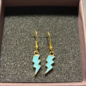 Blixtformade örhängen i guld och turkos - Säljer ett par coola örhängen i form av blixtar. De är i en snygg kombination av guld och turkos, vilket ger dem en unik och trendig look. Perfekta för att liva upp vilken outfit som helst!💙😇