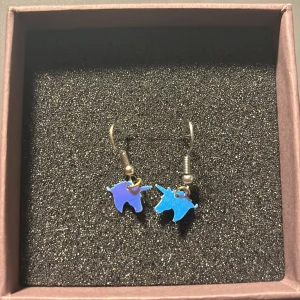 Blå enhörningsörhängen - Säljer ett par supercoola örhängen med små blå enhörningar. De är perfekta för att lägga till en magisk touch till din outfit. Enhörningarna är gjorda av ett glänsande material som fångar ljuset fint. Passar perfekt för både vardag och fest!🦄