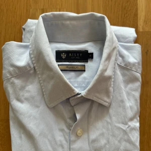 Vit randig skjorta från Riley - Säljer en stilren vit skjorta från Riley med tunna ränder. Skjortan har en klassisk krage och knappar framtill. Perfekt för både vardag och fest! Den är långärmad och i ett mjukt bomullsmaterial.