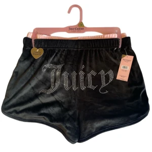 Svarta shorts från Juicy Couture - Säljer ett par snygga svarta shorts från Juicy Couture. De är gjorda i ett mjukt och glansigt material som känns lyxigt. Shortsen har en elastisk midja och en cool Juicy-logga i strass på framsidan. 