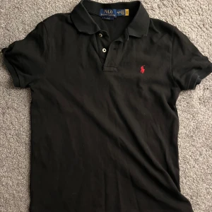 Svart kortärmad pikétröja från Ralph Lauren - Säljer en snygg svart kortärmad pikétröja från Ralph Lauren. Den har en klassisk krage med knappar och rött polo Ralph lauren märke. Använd bara ett par fåtal gånger. Bara att skriva om ni har några frågor!