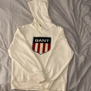 Vit hoodie från GANT - Säljer en snygg vit hoodie från GANT med deras ikoniska logga på bröstet. Den är supermjuk och perfekt för både höst och vinter. Hoodien har långa ärmar och en klassisk passform. Perfekt för en casual look eller att mysa i hemma!
