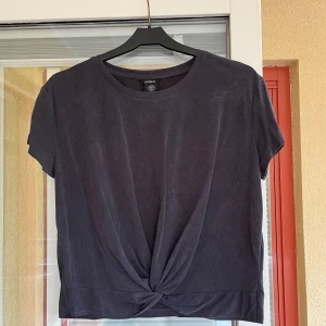 Mörkgrå topp från Lindex - Säljer en mörkgrå topp från Lindex i storlek XS. Den är gjord av en mjuk blandning av modal och polyester med lite elastan för stretch. Toppen har korta ärmar och en knytdetalj framtill som ger den en unik look. Sustainable Choice.