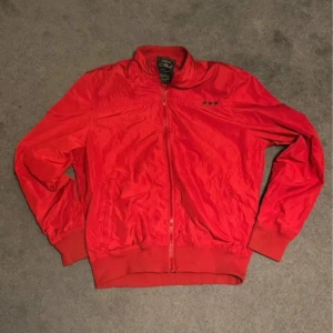 Röd Vindjacka Vår - Väldigt najs fit.  Baggy men sitter bra längst ner pga midjebandet.  Inte regntät men skyddar bra mot vind. Måttligt använd, inga synliga tecken på slitage.  Köp direkt eller lägg prisförslag så kan vi snacka.  Jag skickar vanligtvis inom 1-2 dagar. 
