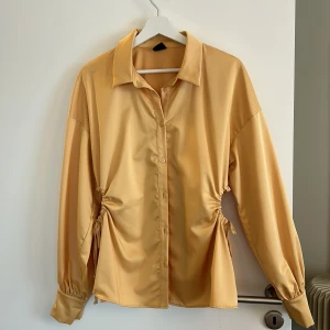 Gul satinblus från Gina Tricot - Säljer en superfin gul blus i satin från Gina Tricot. Den har långa ärmar och knappar framtill. Blusen har en snygg knytning i midjan som ger en cool look. Perfekt för både vardag och fest!