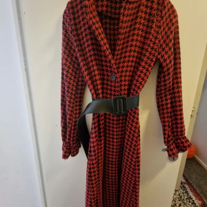 Röd och svart långkappa med bälte - Säljer en supercool röd och svart långkappa med ett klassiskt hundtandsmönster. Kappan har långa ärmar och knäpps med knappar framtill. Den har också ett snyggt bälte i midjan för en mer definierad siluett. Perfekt för höst och vinter!