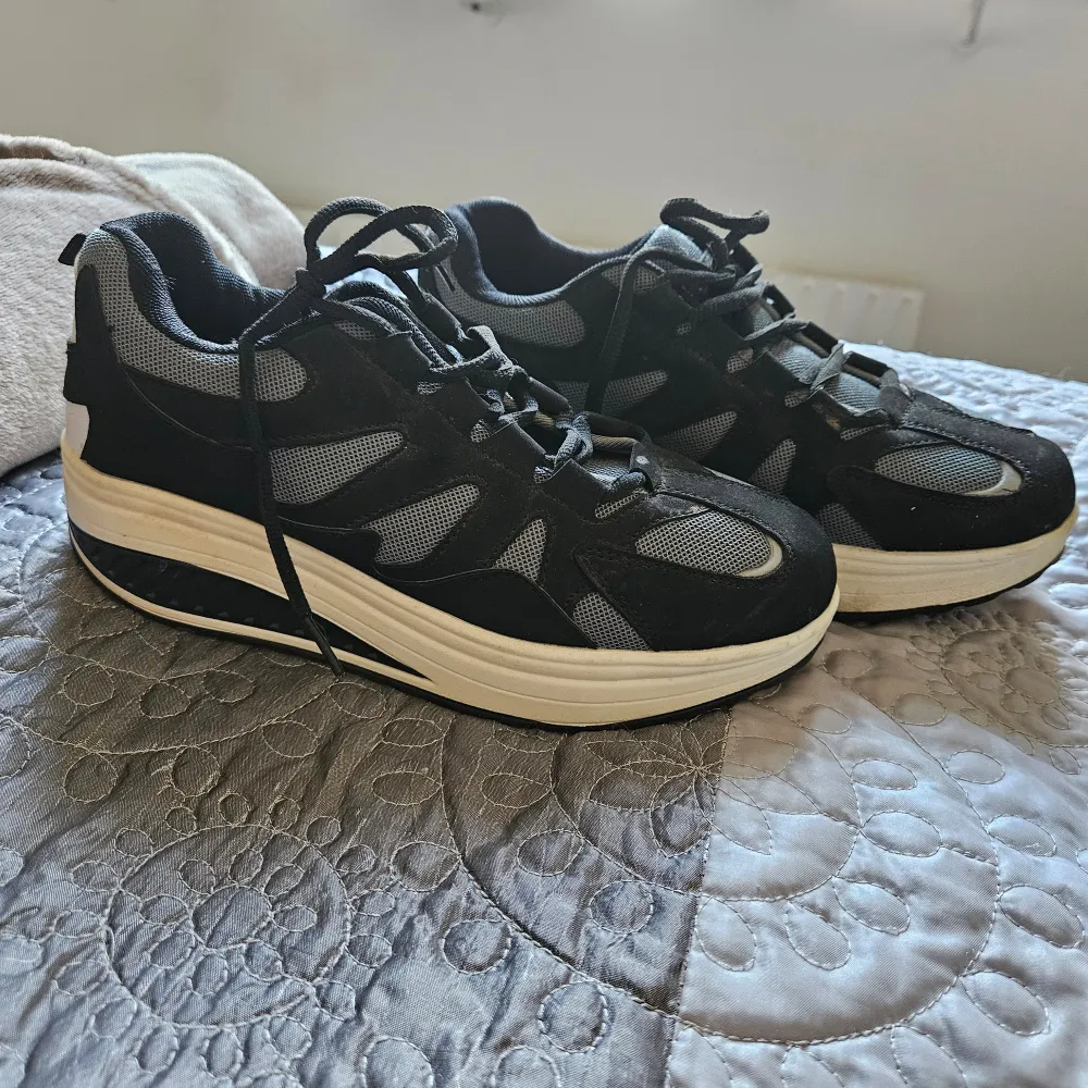 Säljer ett par svart/gråa sneakers med vit sula. De har en sportig design med snörning och är i ett bra skick. Skorna har svarta och grå detaljer och ser ut att vara bekväma för vardagsbruk.. Skor.