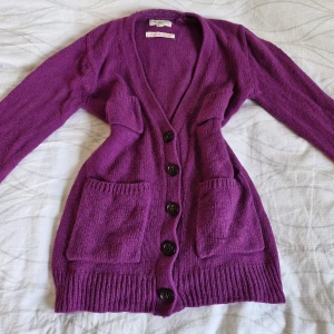 Lila kofta från Holly & Whyte - Säljer en superfin lila kofta från Holly & Whyte by Lindex. Den är stickad och har en v-ringad design med stora knappar framtill. Koftan har långa ärmar och praktiska fickor på sidorna. Perfekt för höst och vinter när du vill hålla dig varm och stilren!