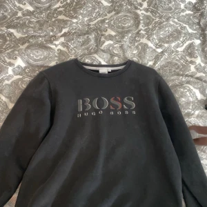 Svart tröja från Hugo Boss - Säljer en snygg svart tröja från Hugo Boss. Den har en klassisk design med märkesloggan tryckt på framsidan. Perfekt för en stilren och avslappnad look. Passar bra till både vardag och fest!