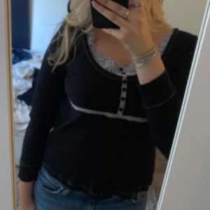 Svart ribbad tröja från Ze-Ze - Säljer en svart ribbad tröja från Ze-Ze i storlek XL men passar oxå L/M. Den har långa ärmar och är dekorerad med vita spetsdetaljer vid halsringningen och längs knappraden. Tröjan har en rad med knappar framtill och är perfekt för en stilren look. 