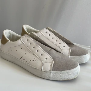 Vita och grå sneakers med guld detaljer - Säljer ett par stilrena sneakers i vitt och grått med snygga gulddetaljer på hälen. Skorna har en perforerad stjärna på sidan och kommer med gråa skosnören. Perfekta för en casual look eller som ett billigare alternativ till golden goose💙