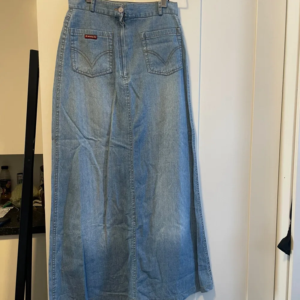 Säljer en snygg lång jeanskjol. Kjolen har en klassisk blå denimfärg och är i ett slitstarkt material. Den har två framfickor med dekorativa sömmar och en knappstängning fram. Perfekt för en avslappnad vardagslook eller att klä upp med en snygg kjol.. Kjolar.