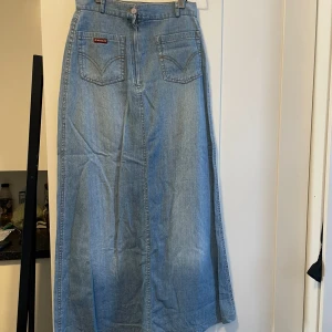 Lång jeanskjol från Rankay - Säljer en snygg lång jeanskjol. Kjolen har en klassisk blå denimfärg och är i ett slitstarkt material. Den har två framfickor med dekorativa sömmar och en knappstängning fram. Perfekt för en avslappnad vardagslook eller att klä upp med en snygg kjol.