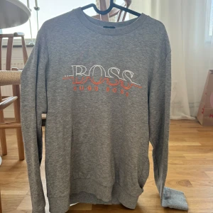 Grå tröja från Hugo Boss - Säljer en snygg grå tröja från Hugo Boss. Den har ett  tryck med 'BOSS' i vitt och 'HUGO BOSS' i orange på framsidan. Tröjan är långärmad och lite tunnare sweatshirt.  Fåtal använd 