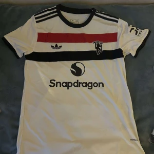 Vit fotbollströja med ränder från Adidas - Säljer en snygg vit fotbollströja från Adidas med coola svarta och röda ränder över bröstet. Tröjan har korta ärmar och en svart krage. Den har också en Snapdragon-logga på framsidan och ett stilrent märke på ärmen. Perfekt för fotbollsfans eller som en casual streetwear-look!