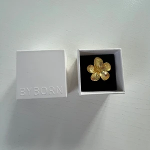 Guldfärgad Blomring från Byborn - Ny justerbar ring   Ny, oanvänd ring i 18k guldpläterad mässing  från Byborn. Modell: Large Flower Ring. Ringen är justerbar. Blomman är 28 mm bred. Nypris: 649 kr.  Elegant guldfärgad ring formad som en blomma från Byborn. Perfekt för att ge en touch av glamour till din outfit. Kommer i en stilren vit ask.