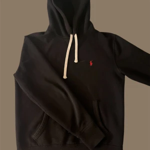 Svart hoodie från Polo Ralph Lauren - Säljer en stilren svart hoodie från Polo Ralph Lauren med klassisk logga på bröstet. Den har en justerbar huva med vita snören och en praktisk magficka. Perfekt för en avslappnad look.