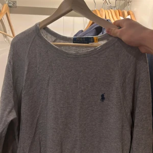 Grå sweatshirt från Polo Ralph Lauren - Säljer en stilren grå tröja från Polo Ralph Lauren med det klassiska logotypbroderiet i blått på bröstet. Tröjan har en rund halsringning och långa ärmar. Tröjan är endast ändats använd 1 gång så den är i bra skick