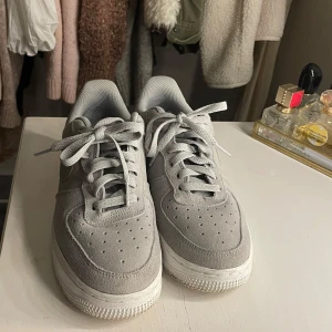 Grå Nike Air Force sneakers - Snygga grå Nike Air Force sneakers i mocka med klassisk design och vit sula. Skorna har snörning och en subtil gulddetalj på hälen. Perfekta för en stilren look.