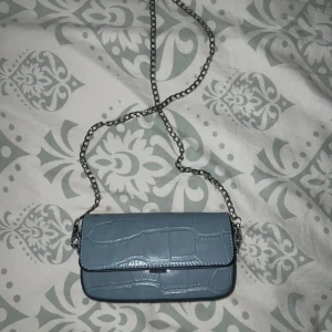 Blå axelväska med kedja - Snygg blå axelväska med krokodilpräglat mönster och en stilren kedjerem. Väskan har en praktisk storlek och ett enkelt spänne för att hålla dina saker säkra. Perfekt för att ge din outfit en trendig touch.