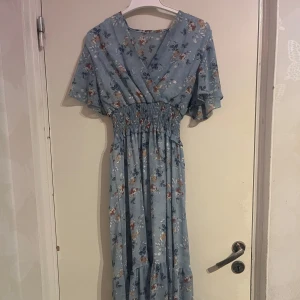 Blommig blå långklänning - Charmig blå långklänning med blommigt mönster. Klänningen har en omlott-design och kortärmade ärmar. Perfekt för sommarens alla tillfällen.