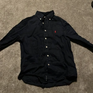 Polo ralph lauren mörkblå linneskjorta knappt använd !! Priset ej hugget i sten!! - En stilren mörkblå linneskjorta från Polo Ralph Lauren, perfekt för sommaren, är knappt använd, Storlek 10/12 men passar även XS. OBS!! PRISET EJ HUGGET I STEN!! Säljer för att den är för liten och knappt kommit till användning 