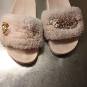 Ljusrosa tofflor med päls och gulddetaljer - Snygga ljusrosa tofflor med päls och glittrande gulddetaljer. Perfekta för en lyxig och bekväm stil. Sandalerna har en bred rem över foten med en dekorativ kedja och en liten gulddetalj på sidan, pga att den har gått sönder. 