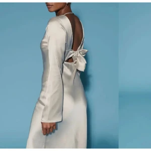 Beige satin klänning från Gina Tricot - Elegant beige klänning i satin från Gina Tricot. Klänningen har en öppen rygg med en stor rosett som detalj och långa ärmar. 