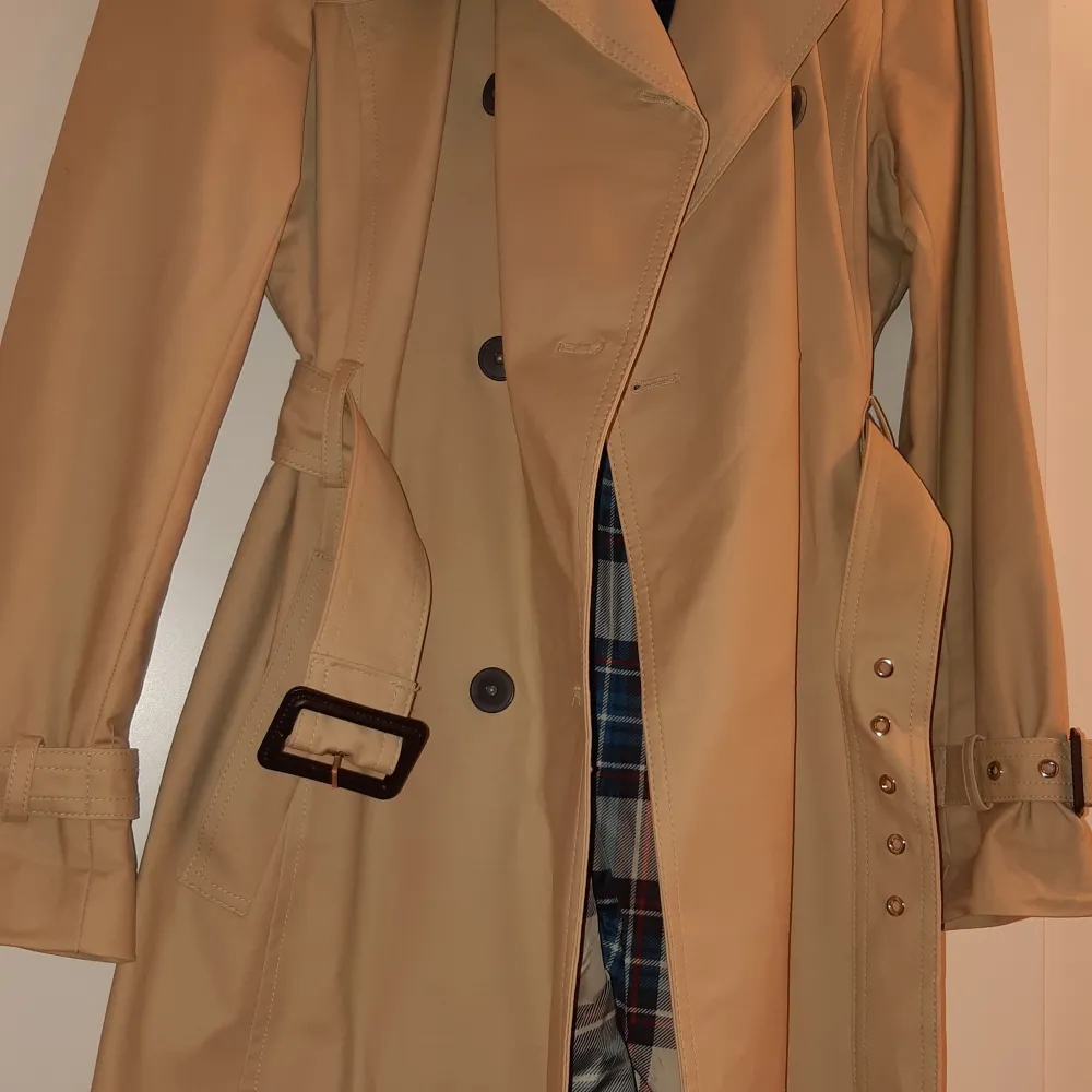 Snygg beige trenchcoat från Mango aldrig använt, med klassisk dubbelknäppning och bälte i midjan. Jackan har långa ärmar och ett stilrent rutigt foder. Perfekt för höst och vår!. Takit.