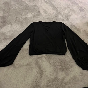 Svart omlottopp från Gina Tricot - Snygg svart omlottopp från Gina Tricot i polyester med en elegant v-ringning. Toppen har långa pösiga ärmar och en stilren design som passar perfekt för olika tillfällen.