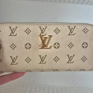 Snygg beige plånbok med det klassiska monogrammönstret i brunt. Plånboken har en dragkedja i guld och är perfekt för att hålla dina kort och kontanter säkra. En stilren accessoar för alla tillfällen.