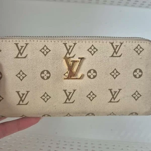 Beige plånbok  - Snygg beige plånbok med det klassiska monogrammönstret i brunt. Plånboken har en dragkedja i guld och är perfekt för att hålla dina kort och kontanter säkra. En stilren accessoar för alla tillfällen.