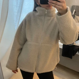 Beige teddyjacka med dragkedja - Mysig beige teddyjacka med dragkedja och hög krage. Perfekt för kyliga dagar när du vill hålla dig varm och bekväm. Jackan har en avslappnad passform och långa ärmar. Oversized.