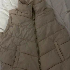 Beige dunväst från JDY. Knappt använd. - Säljer en snygg beige dunväst från JDY. Västen har en dragkedja framtill och är perfekt för kyligare dagar. Västen håller dig väldigt varm. Knappt använd inga fläckar/ i helt skick. Köpt från Zalando men säljer pga fel storlek.