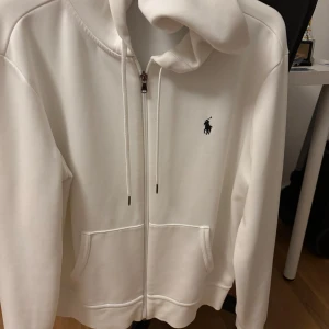 Vit hoodie från Ralph Lauren - Säljer en stilren vit hoodie från Ralph Lauren med dragkedja och klassisk logga på bröstet. Tröjan har en justerbar huva med snören och praktiska fickor framtill. Perfekt för en avslappnad look. Storlek M passar även S