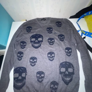 Grå tröja med dödskallemönster från Philipp Plein - Cool grå tröja från Philipp Plein med flera svarta dödskallemotiv över hela plagget. Tröjan har rund halsringning och är tillverkad i mjukt stickat material. Metallmärke med logga på bröstet ger extra edge till looken.