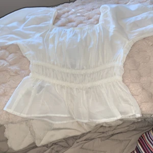 Vit blus från H&M - Säljer en söt vit blus från H&M med elastiska detaljer i midjan och vid ärmsluten. Blusen har en luftig passform och trekvartsärmar, perfekt för vår och sommar. Den är tillverkad i ett lätt matrial. Oanvänd!!!