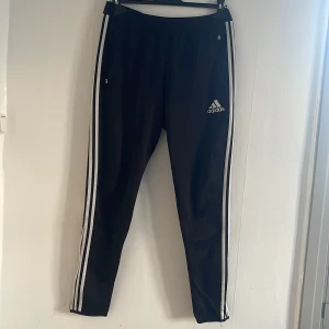 Svarta träningsbyxor från Adidas - Snygga svarta träningsbyxor från Adidas med vita ränder längs sidorna och Climacool-teknologi för optimal ventilation. Byxorna har en elastisk midja och dragkedjor vid anklarna för enkel på- och avtagning.