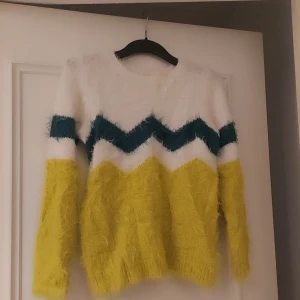 Flerfärgad fluffig  tröja  till flickor från H&M, storlek 10 år - Säljer en mysig och fluffig stickad tröja till flicka från H&M i vitt, blått och gult med ett zigzag-mönster. Tröjan har långa ärmar och är perfekt för att hålla sig varm under kyliga dagar. Den är både stilren och bekväm.