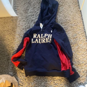 Mörkblå hoodie från Ralph Lauren - Säljer en snygg mörkblå hoodie från Ralph Lauren med röd och vit detaljering. Tröjan har en klassisk passform med huva och långa ärmar. Perfekt för en avslappnad stil. 🧢