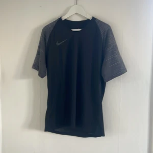 Svart Dri-FIT t-shirt från Nike - Säljer en svart Dri-FIT t-shirt från Nike med gråmelerade ärmar. Perfekt för träning med sin fukttransporterande teknologi. T-shirten har en normal passform och korta ärmar. Nike-loggan är diskret placerad på bröstet.
