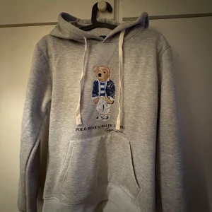 Grå hoodie med Polo Bear från Ralph Lauren - Säljer en grå hoodie från Ralph Lauren med den ikoniska Polo Bear på framsidan. Tröjan har en känguruficka och justerbar huva med snören. Perfekt för en avslappnad stil.