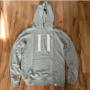 Grå les deux hoodie - Säljer en stilren grå hoodie med ett stort vitt tryck av två romerska siffror på framsidan. Tröjan har en klassisk design med huva och en stor ficka framtill. Perfekt för en avslappnad stil.