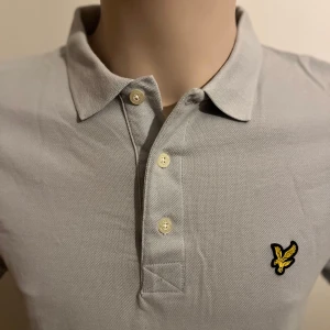 Grå pikétröja från Lyle & Scott - Säljer en stilren grå pikétröja från Lyle & Scott med klassisk krage och knappslå. Tröjan har en broderad logotyp i gult och svart på bröstet. Perfekt för en avslappnad och snygg look.