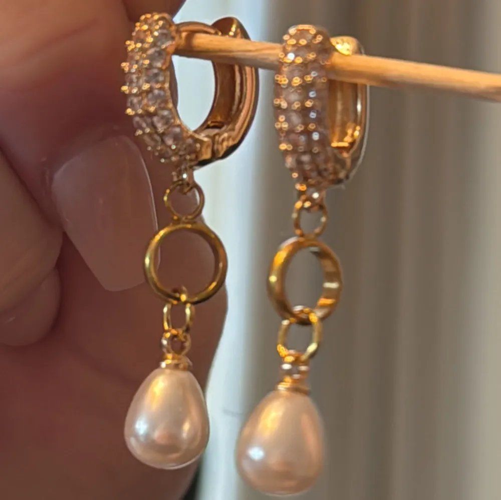 Eleganta guldörhängen med glittrande strass och hängande pärlor. Perfekta för att ge en lyxig touch till din outfit. De har en cirkulär design med två ringar och en pärla som hänger längst ner.. Asusteet.