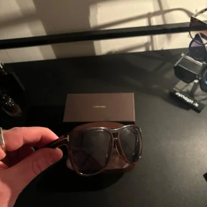 Solglasögon från Tom Ford - Snygga solglasögon från Tom Ford med en elegant brun båge och mörka linser. Perfekt för att ge en stilren touch till din look. Kommer med ett matchande fodral för säker förvaring.