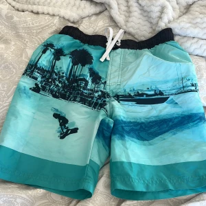 Turkosa badshorts med strandmotiv - Snygga turkosa badshorts med ett coolt strandmotiv i blå nyanser. De har en elastisk midja med vit snörning för justerbar passform. Perfekta för sommarens strandhäng eller poolpartyn! Jag tror st är 146 då jag jämfört med ett annat par shorts!! Lappen är tyvärr ej kvar i