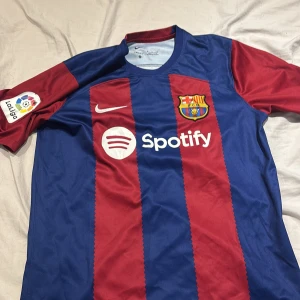Barcelona kit  - Säljer en blå och röd randig fotbollströja från Nike med korta ärmar. Tröjan har klubbmärke och sponsorloggor på framsidan samt UNHCR-logga på baksidan. Perfekt för fotbollsfans!