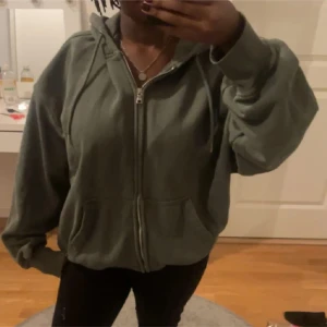 Grön hoodie med dragkedja - Säljer en mysig grön hoodie med dragkedja framtill. Den har en avslappnad passform och långa ärmar, perfekt för kyliga dagar. Huvan ger extra värme och stil. Passar bra till både jeans och träningsbyxor.