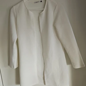 Vit kavaj från ONLY - Snygg vit kavaj från ONLY i en stilren design. Kavajen har en öppen front utan knappar och är tillverkad i ett lätt material. Perfekt för att ge en elegant touch till din outfit.